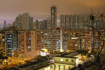 Fototapeta premium Cityscape of Sham Shui Po skyline in Kowloon, Hong Kong with brightly lit skyscraper buildings, light pollution in sky and silhouetted tree in foreground