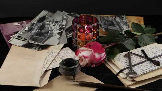 Burning candle in a glass candlestick, a pink rose and falling petals on the background of old letters and photographs. Family archive, memory