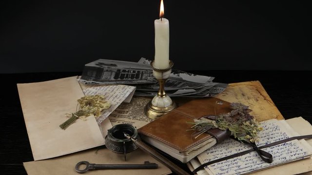 A burning candle, dried flowers, an old key, a writing-pad and an inkwell with a pen on the background of old letters and photographs. Memory, family archive