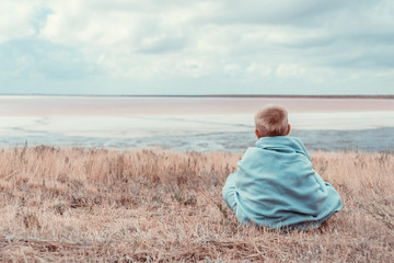Naklejka na ściany i meble Small boy is sitting near the sea wreapped in warm blanket