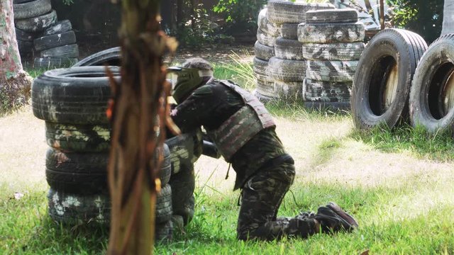 Man in Camouflage Suit with Gun and Protective Mask Playing Paintball Game on Paintball Field with Tires and Wooden Shelters in Forest. Extreme Team Sport