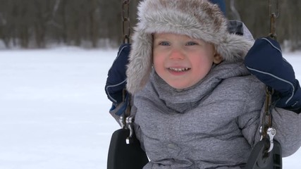kid swaying on swing. Cute little boy (toddler, two year old) in hat with fur trim and earflaps.  Happy child. Winter, cold weather, outdoor