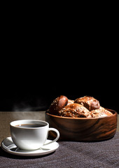 A cup with a hot drink and a wooden plate with pies stand on a linen tablecloth against a black background.
