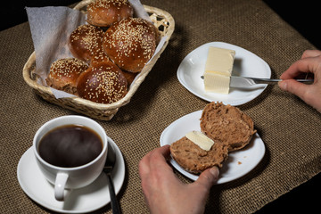 Female hand puts butter on a sliced sesame bun. A cup of hot drink stands nearby.