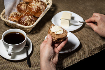 Female hand puts butter on a sliced sesame bun. A cup of hot drink stands nearby.