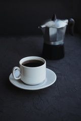 White cup of coffee and coffee maker on dark background