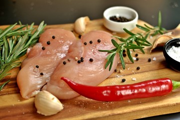 Raw fresh uncooked chicken breast meat fillet dish with rosemary, pepper, salt and red hot chili pepper on wooden board and black background. Front side view