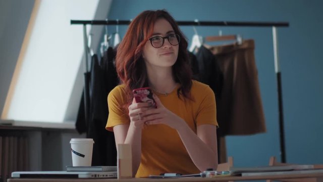 A stylish young woman in a bright T-shirt is sitting at the table, looking at the smartphone screen is reflected in the glasses. Her colleague moves the coat hanger and outweighs the dresses.