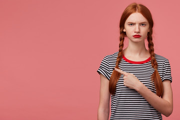 Serious displeased angry girl with two red haired braids red lip , dressed in stripped t-shirt, pointing fore finger to the upper left corner at blank copy space isolated on pink background