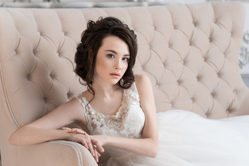 Beautiful bride sitting on the couch. Wedding hairstyle and makeup.  Lace dress. The hand on the back of the sofa. Beige sofa.