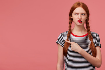 Doubtful thoughtful girl with two red haired braids biting red lip thinks about something, dressed in stripped t-shirt, looks to the upper left corner pointing fore finger isolated on pink background