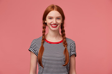 Nice pretty red-haired girl with red lips, two braids, chrarming smile, shows white healthy teeth, dressed in stripped t-shirt, isolated on a pink background