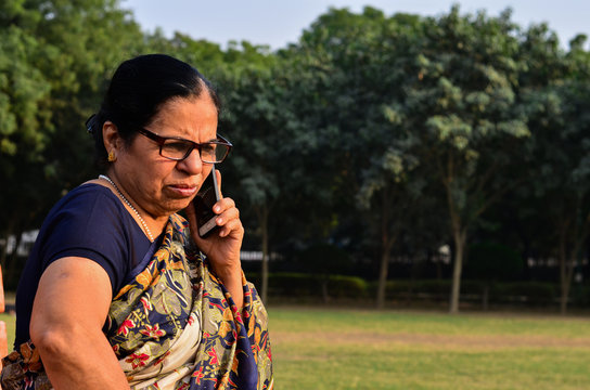 Senior Indian Woman Giving Angry Expressions Speaking On Her Smartphone, Sitting On A Bench In A Park In Delhi. Concept Shot Showcasing Technology Adoption By Senior Citizens. Digital India