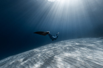 Woman freediver glides in the depth among the school of fish over the sandy bottom with sun rays...