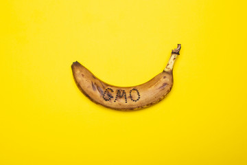 Overripe, blackened banana on whose skin the word “GMO” is written lies on a yellow background.