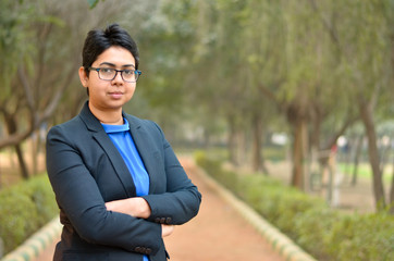 Closeup portrait of a confident young Indian Corporate professional woman with short hair and spectacles, crossed folded hands in an outdoor setting wearing a black business / formal suit 