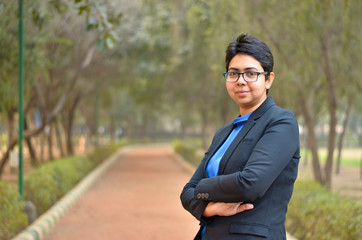 Closeup portrait of a confident young Indian Corporate professional woman with short hair and spectacles, crossed folded hands in an outdoor setting wearing a black business / formal suit 