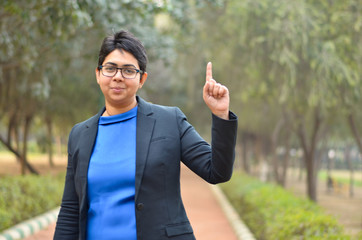 Portrait of a confident young Indian Corporate professional woman with short hair pointing upwards with her finger in an outdoor setting wearing a black business / formal suit 