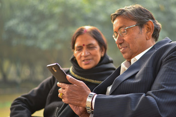 Side portrait of a senior Indian couple sitting in park looking at their smart phone in Delhi