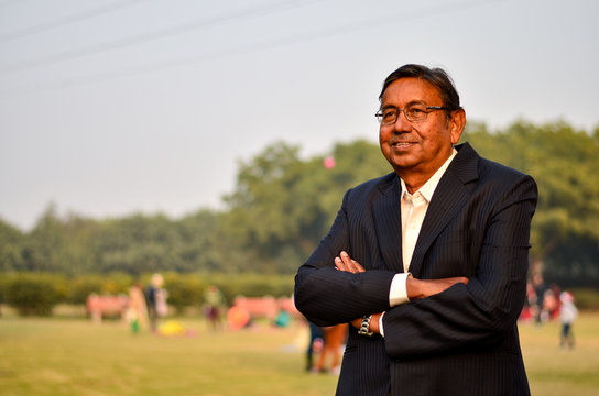 Portrait Of A Happy Senior Indian Man Wearing A Suit In The Outside Setting Standing With Crossed Hands And Thinking In A Park In Delhi, India