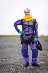 Pretty woman happy, dressed in jumpsuit and parachute harness on airport runway ready for skydive jump