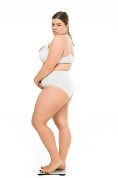 side view of sad young overweight woman in underwear standing on scales isolated on white, lose weight concept