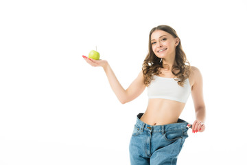 smiling slim young woman in big jeans holding green apple isolated on white