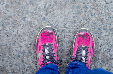 This is a top view of pink colored sports shoes with tied laces. the shoes are worn by a girl woman standing on a tarmac road also wearing blue jeans or trousers