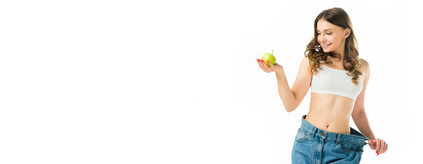smiling slim young woman in big jeans holding ripe green apple isolated on white