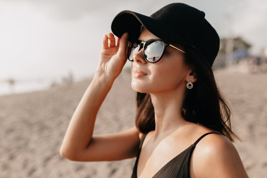 Stylish modern woman in trend outfit looking on the ocean with happy smile wearing cap and glasses and enjoys summer hot days.  Young caucasian female model on the sea shore