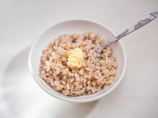 Barley porridge with a piece of butter in a deep white plate with a spoon on a white background