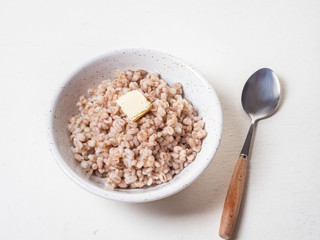 Barley porridge with a piece of butter in a deep white ceramic plate, next to it lies with a spoon with a wooden handle on a white background, shot close-up