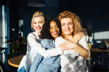 Portrait of three multiethnic women teenagers friends laughing and hugging while spending time together indoor over cafe interior background. Multi-racial and difference of cultures friendship concept