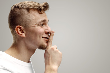 Keep silence. Handsome young man in white shirt looking at camera and holding finger on lips while standing against grey background