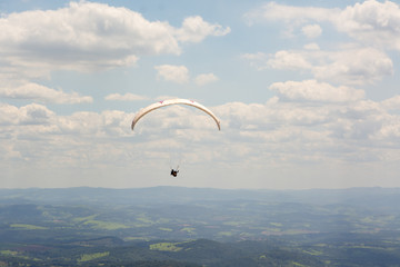 Lone paraglider gliding through the air over a green, mountainous backdrop and partially clouded skies.