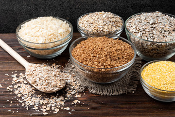Carbohydrates. Various grain flakes in bowls on dark wooden background. Oat, corn, rice, buckwheat, rye and wheat flakes.