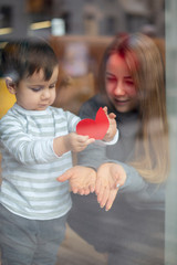Mom and little boy are having fun on a sunny day by the window in a cafe, holding a red paper heart, a symbol of love. Happy mother's day. selective focus, noise effect