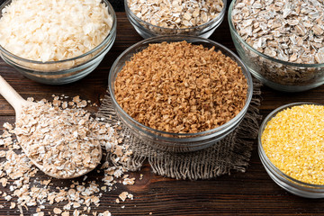 Carbohydrates. Various grain flakes in bowls on dark wooden background. Oat, corn, rice, buckwheat, rye and wheat flakes.