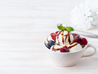 Balls of vanilla ice cream in mug with raspberry and blueberries, chocolate syrup  topping. Side view, copy space, white bright background