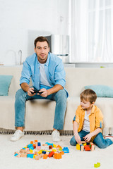 handsome father with joystick playing video game while cute preschooler son playing with colorful building blocks at home