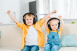 adorable brothers in headphones looking at camera and cheering with hands in air at home