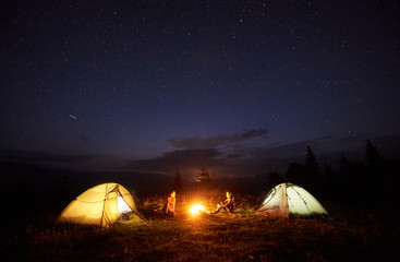 Fototapeta na wymiar Camping in mountains in the evening. Bright bonfire burning between two tourists, man and woman sitting opposite each other near illuminated tents under night starry sky on distant hills background.