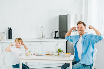 smiling preschooler son and father looking at camera, posing and showing biceps during breakfast in kitchen