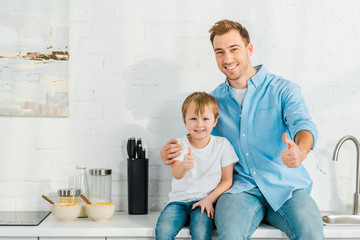 father and preschooler son looking at camera and showing thumbs up signs during breakfast in kitchen