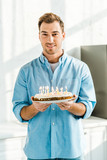 handsome man looking at camera and holding birthday cake with burning candles at home in morning