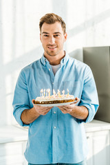 handsome man looking at camera and holding birthday cake with burning candles at home in morning