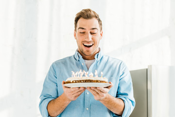 excited handsome man holding birthday cake with burning candles at home in morning