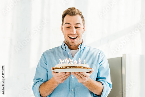 excited handsome man holding birthday cake with burning candles at home in morning