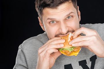  man eating tasty chicken burger while looking at camera isolated on black