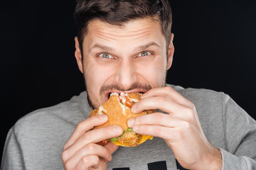 handsome man eating tasty chicken burger while looking at camera isolated on black © LIGHTFIELD STUDIOS